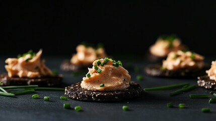 Salmon mousse elegantly piped onto dark rye crackers, arranged against a black background with contrasting chives