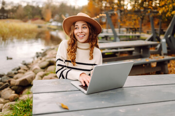 Working surrounded by autumn nature. Young woman with laptop at a rustic table near a tranquil waterway in autumn. E-learning concept. Remote work.