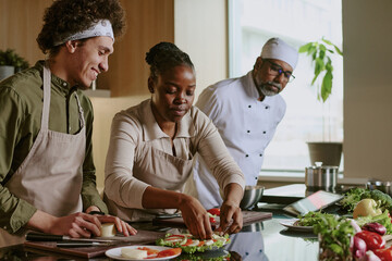 African American female cook decorating caprese appetizer while curly haired sous chef and Indian chef watching her actions