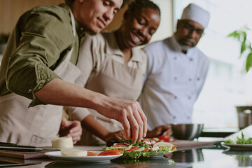 Medium close up of young sous chef adding condiment into caprese appetizer while female cook and chef looking at him