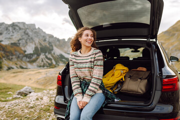 Young woman sits in the trunk of her car, parked on the side of a winding road that cuts through...