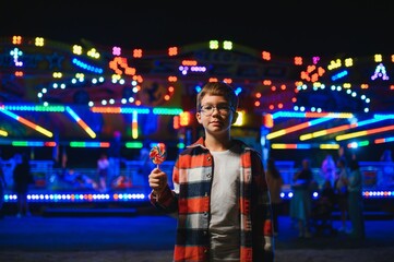 A cute caucasian boy eating a colorful lollipop standing against the background of a carousel in...
