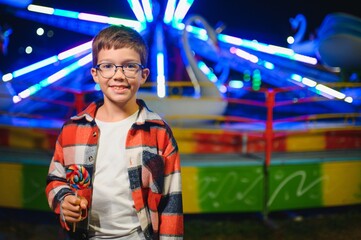 A cute caucasian boy eating a colorful lollipop standing against the background of a carousel in...