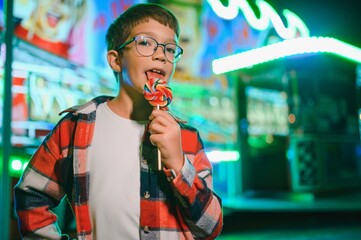 A cute caucasian boy eating a colorful lollipop standing against the background of a carousel in...