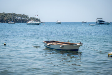 A small wooden boat anchored in calm waters surrounded by larger boats on a sunny day near a serene...