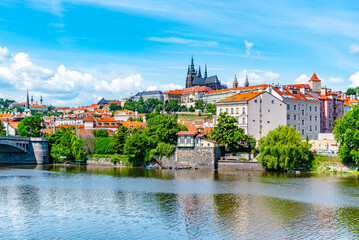 A vibrant view capturing the historic Prague Castle and Manes Bridge on a sunny day with clear skies, overlooking the calm Vltava River.