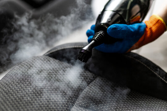 Steam cleaning a car seat using a handheld cleaner in a garage setting during daylight hours