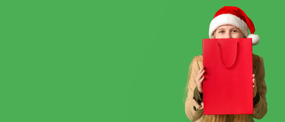 Beautiful young woman in warm winter clothes and Santa hat with shopping bag on green background