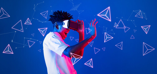 Smart boy standing at blue background while floating geometric shape. African man playing game and wearing VR goggle and casual shirt. Augmented reality technology innovation concept. Ingenuity.