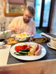 hearty breakfast is beautifully arranged on a plate, featuring sausages, fried eggs, and fresh vegetables. in the background, a person prepares to enjoy this appetizing meal, highlighting a cozy