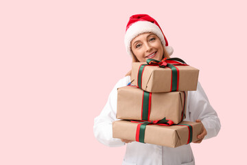 Mature female doctor in Santa hat with Christmas gift boxes on pink background