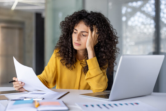 Hispanic businesswoman sits at desk with laptop, reviewing documents with concentrated expression. Image conveys stress, analysis, and multitasking in workplace setting.