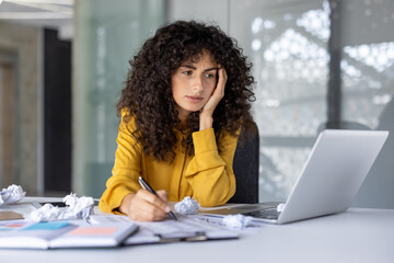 Hispanic businesswoman experiencing stress at work desk using laptop surrounded by crumpled paper...