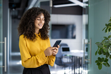 Hispanic businesswoman smiling as she uses smartphone in modern office. Scene conveys technology, communication, success, and professional atmosphere, highlighting effective use of digital tools