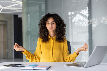 Young woman meditating at office desk with closed eyes. Practicing mindfulness and relaxation for stress relief during work. Laptop, notebook nearby, promoting calm and focus in busy environment.
