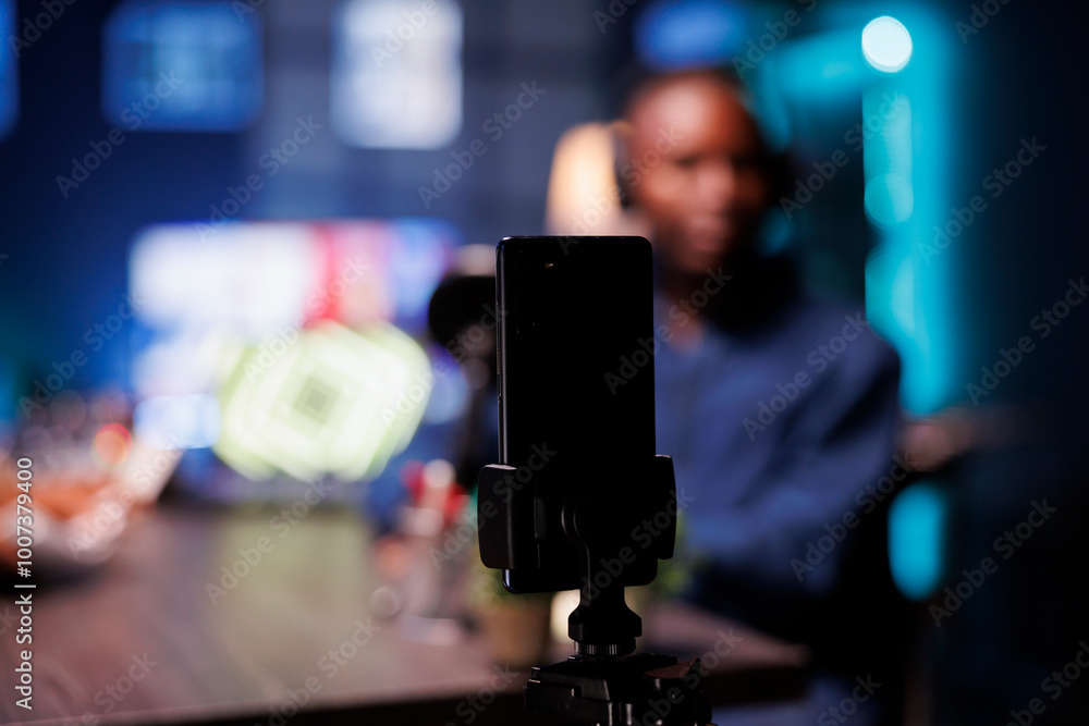 Poster focus on smartphone on tripod recording vlogger show host in blurry background interviewing guest in