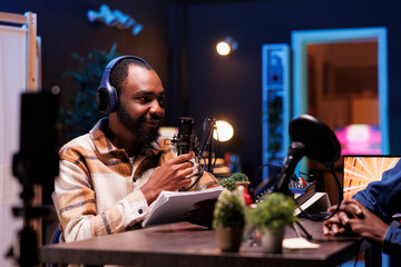 Journalist is recording an interview with his cellphone mounted on a tripod. He reviews notes for relevant questions and films a vlog with his mobile device while talking to celebrity on microphone.