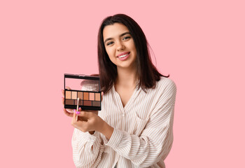 Beautiful young woman with palette of eyeshadows and makeup brush on pink background