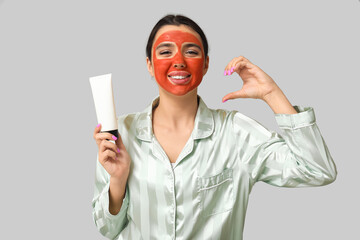 Beautiful young woman with facial mask making half of heart on grey background