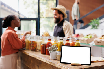 Tablet with a blank chromakey mockup template horizontally positioned near glass jars. On the counter is a smart device displaying an isolated white screen while shopkeeper converses with customer.