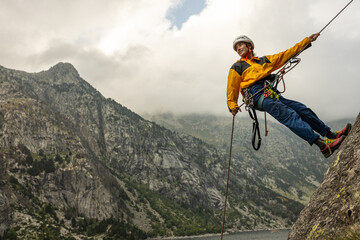 A man is hanging from a rope on a mountain. The man is wearing a yellow jacket and blue pants. The mountain is covered in trees and the sky is cloudy