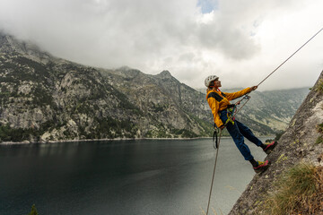 Obraz premium A man is climbing a rock face with a rope attached to him. The scene is set against a backdrop of mountains and a lake. The man is wearing a yellow jacket and he is enjoying the challenge of the climb