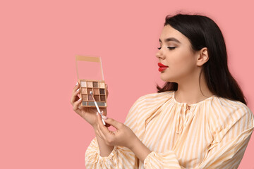 Beautiful young woman with palette of eyeshadows and makeup brush on pink background