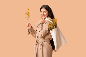 Happy young woman holding eco bag with mimosa flowers on beige background