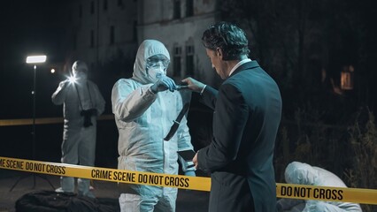 Detective examining main clue brought in by forensic scientist at crime scene, while the rest of the experts collect remaining evidence behind police tape next to headless body. Night scene.