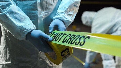 A forensic scientist rolling out police tape at a crime scene at night, while other experts collect...