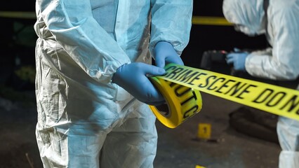 A forensic scientist rolling out police tape at a crime scene at night, while other experts collect evidence in the background