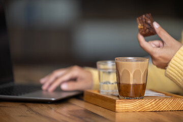 Close-up happy young businesswoman self-employed woman enjoying a caramel coffee and brouni in a specialized coffee shop, works with her laptop, digital nomad, enjoys a coffee on a wooden table