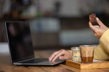Freelance young happy woman enjoying a delicious breakfast of caramel coffee and brouni in a specialized cafe, works with her laptop, digital nomad, thoughtfully enjoys a coffee on a wooden table