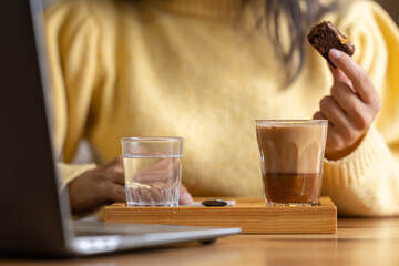 Close-up happy young businesswoman self-employed woman enjoying a caramel coffee and brouni in a specialized coffee shop, works with her laptop, digital nomad, enjoys a coffee on a wooden table