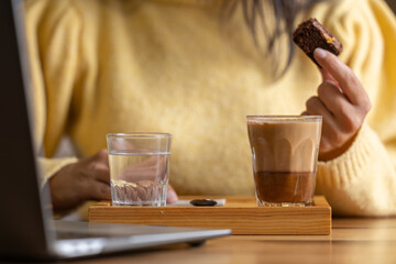 Close-up happy young businesswoman self-employed woman enjoying a caramel coffee and brouni in a specialized coffee shop, works with her laptop, digital nomad, enjoys a coffee on a wooden table