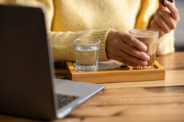 Close-up happy young businesswoman self-employed woman enjoying a caramel coffee and brouni in a specialized coffee shop, works with her laptop, digital nomad, enjoys a coffee on a wooden table