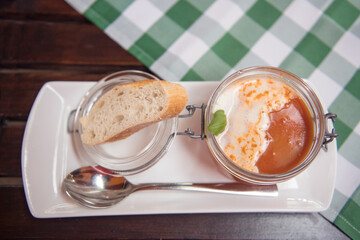 A delicious serving of creamy tomato soup in a glass jar, garnished with basil, accompanied by a slice of fresh bread on a white plate with a spoon, set on a checkered green tablecloth.
