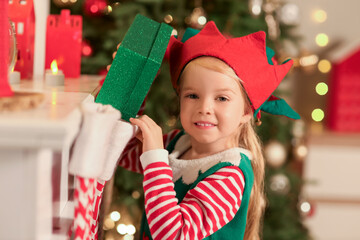 Cute little girl in elf costume taking gift from sock at home on Christmas eve, closeup