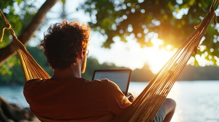 A man sits comfortably in a hammock, using his laptop under a canopy of leaves by the water, with sunlight streaming through the foliage on a peaceful afternoon.