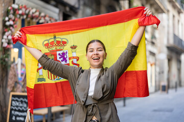 Young woman in casual clothes holding big flag of Spain while standing on street
