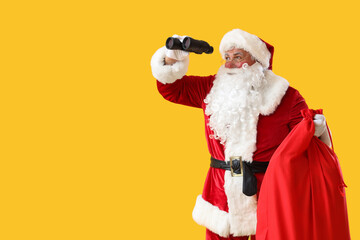 Santa Claus with Christmas bag looking through binoculars on yellow background