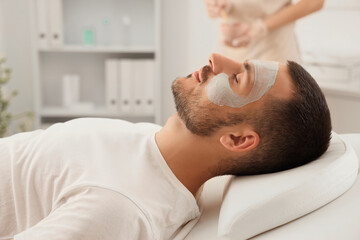 Beautician and handsome young man with cosmetic clay mask on face in spa salon