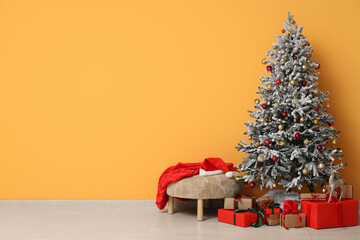 Decorated Christmas tree with gift boxes, pouf with Santa hat and sack near yellow wall