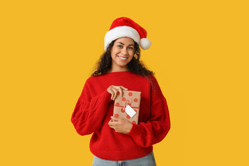 Happy African-American woman in Santa hat and with Christmas gift on yellow background