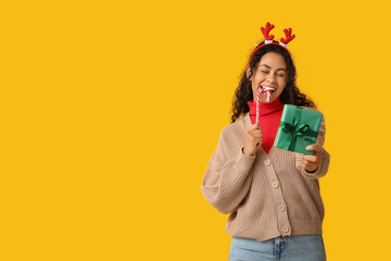 Happy African-American woman with Christmas gift and candy cane on yellow background