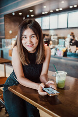 A young woman sits at a table in a modern coffee shop, smiling while using her smartphone and enjoying a green iced drink.