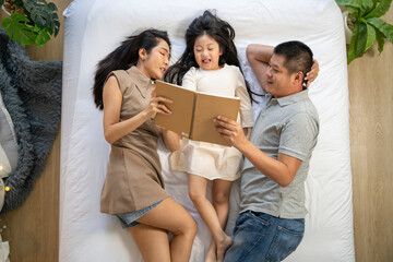 A family enjoys quality time together, reading a book while lying on the bed. The parents and their daughter share a relaxing moment, strengthening their bond in a peaceful home environment.