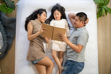Obraz premium A family enjoys quality time together, reading a book while lying on the bed. The parents and their daughter share a relaxing moment, strengthening their bond in a peaceful home environment.