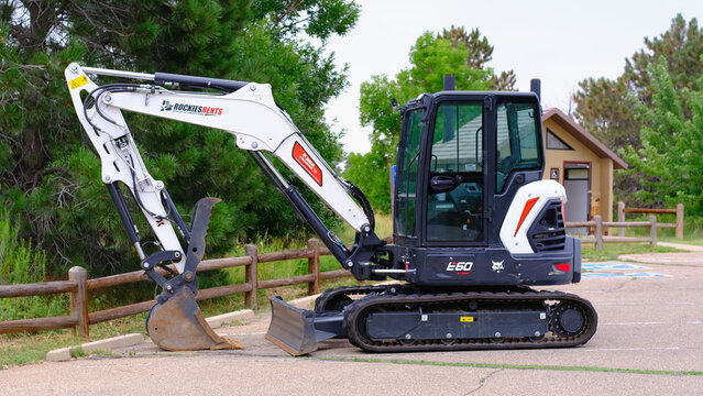 Bobcat mini excavator parked on paved surface at public park restroom building surrounded by green trees and fenced pathway.Construction, public works, heavy equipment rental, landscaping services.