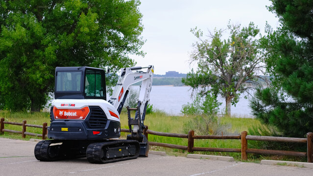 Bobcat compact excavator parked on paved area near a wooden fence overlooking a lake with trees in the background. Construction, public works, heavy equipment rental, landscaping services.